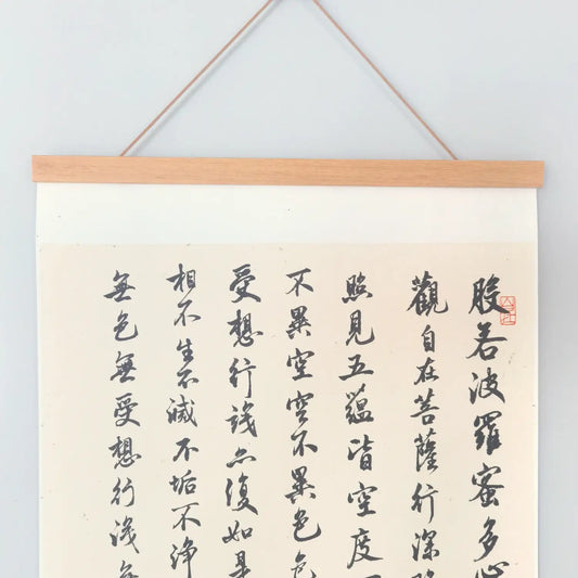 Handwritten Heart Sutra Chinese calligraphy hanging scroll with teak rods, displayed on a minimal white wall.