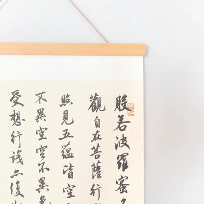 Handwritten Heart Sutra Chinese calligraphy hanging scroll with teak rods, displayed on a minimal white wall.